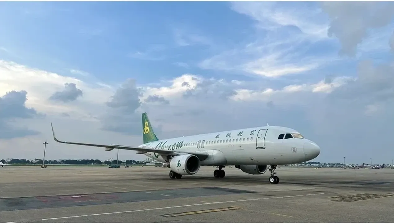 Một chiếc máy bay màu trắng của Hãng hàng không Spring Airlines đậu trên sân đỗ, một chiếc máy bay phản lực hiện đại với thương hiệu màu xanh lá cây phản chiếu sắc thái dịu dàng của bầu trời.